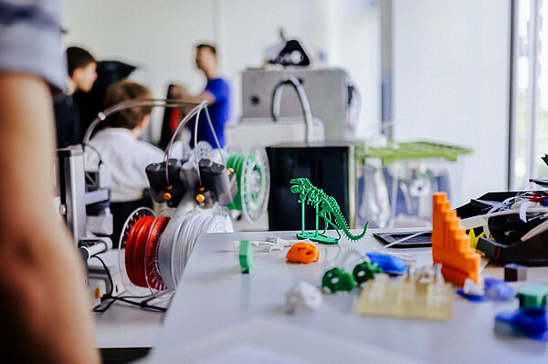 3D-gedruckte Exemplare wie ein kleiner grüner Dino auf dem Tisch, im Hintergrund Dozent unscharf zu sehen